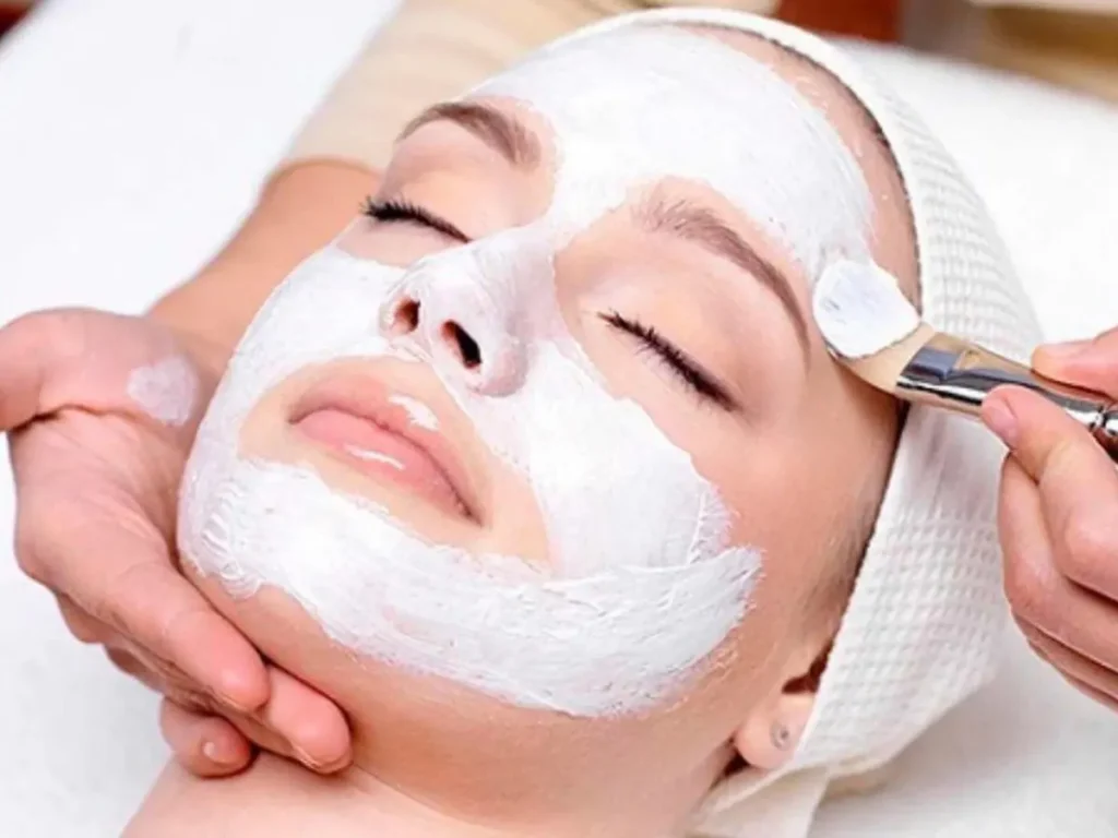 Woman relaxing with eyes closed while an esthetician brushes a facial mask onto her skin