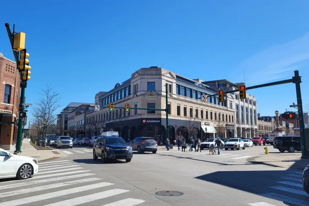 Street corner in downtown Birmingham, Michigan with a multi-story stone retail and office building, crosswalks and traffic signals under a clear blue sky