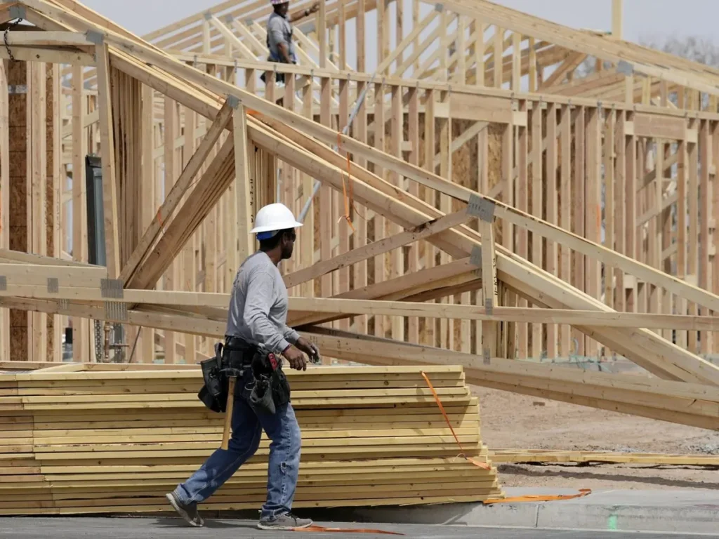 Framing crew in hard hats carrying lumber across a wood-frame house under construction