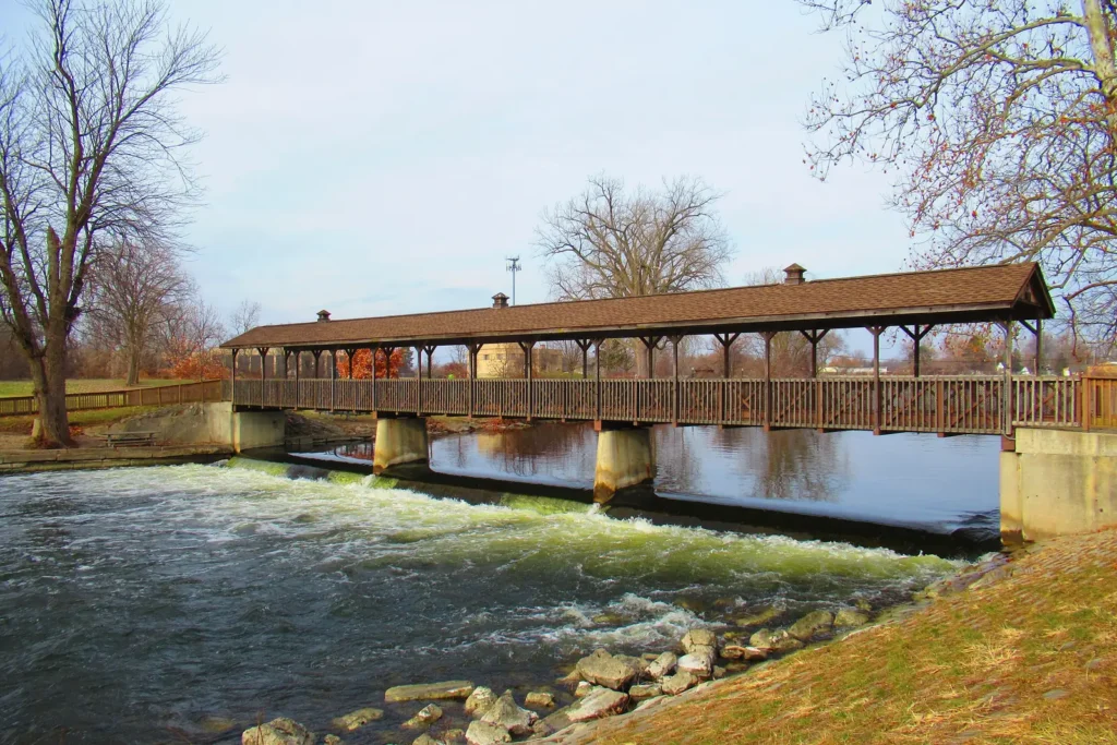 A brown covered wooden footbridge crossing the small Huroc Dam on the Huron River in Flat Rock, with white water spilling over the dam and bare trees on a clear day