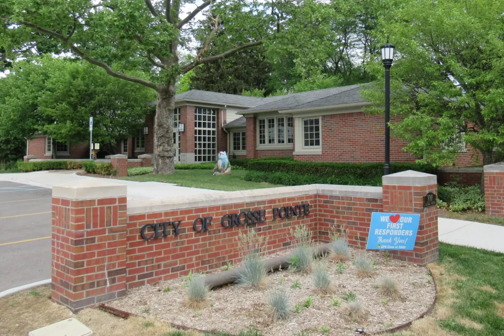 Red brick City of Grosse Pointe municipal building behind a brick sign wall reading City of Grosse Pointe, framed by mature trees on a spring day