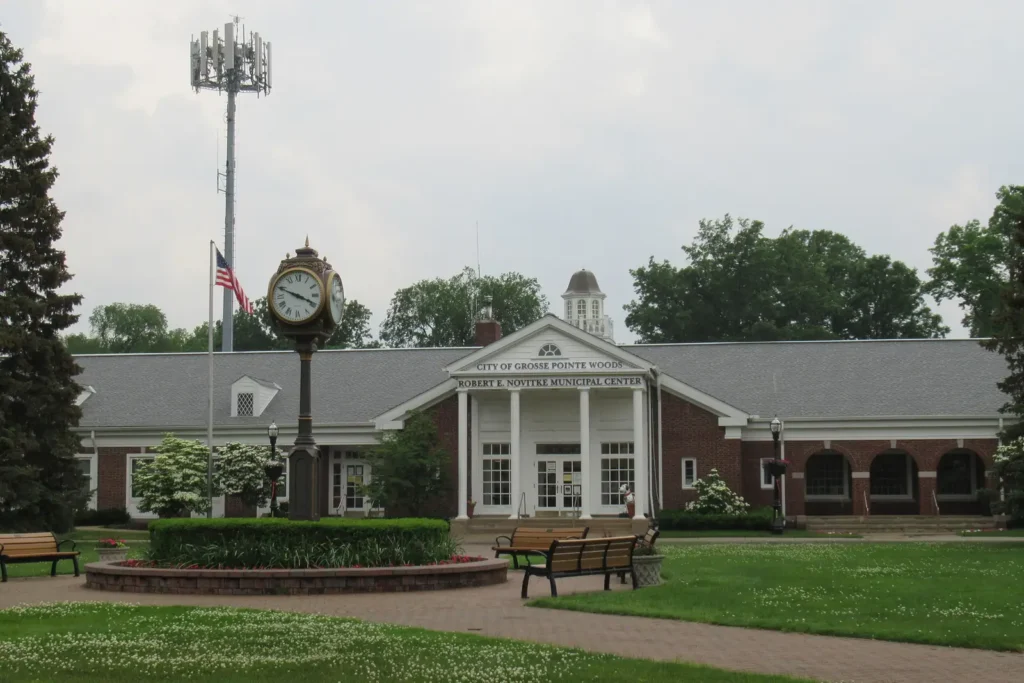 The red brick Robert E. Novitke Municipal Center in Grosse Pointe Woods with a white columned entrance, a post clock and benches on the front lawn