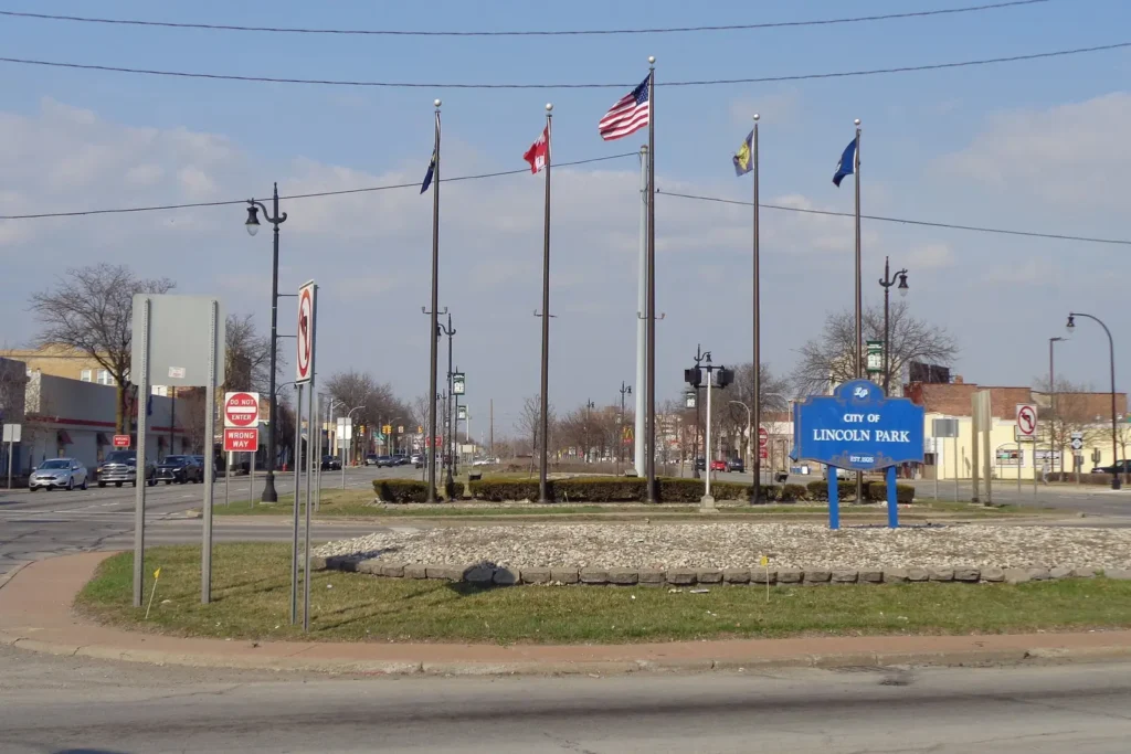 City of Lincoln Park sign and flagpoles in a planted median at Southfield Road and Fort Street, with downtown storefronts behind