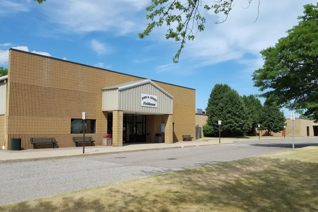 The tan brick John K. Kessey Fieldhouse at the Melvindale Civic Center on a sunny summer day, with trees along the drive