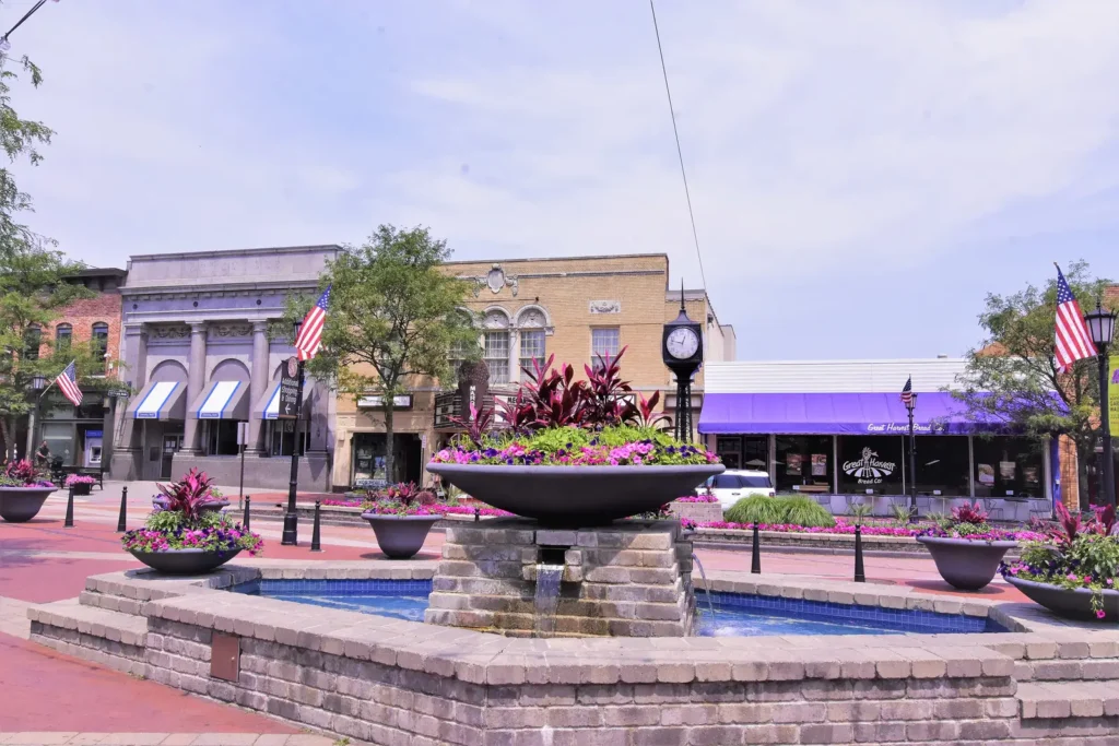 The town square fountain, flower planters, street clock and storefronts on Main Street in downtown Northville decorated with American flags on a summer day