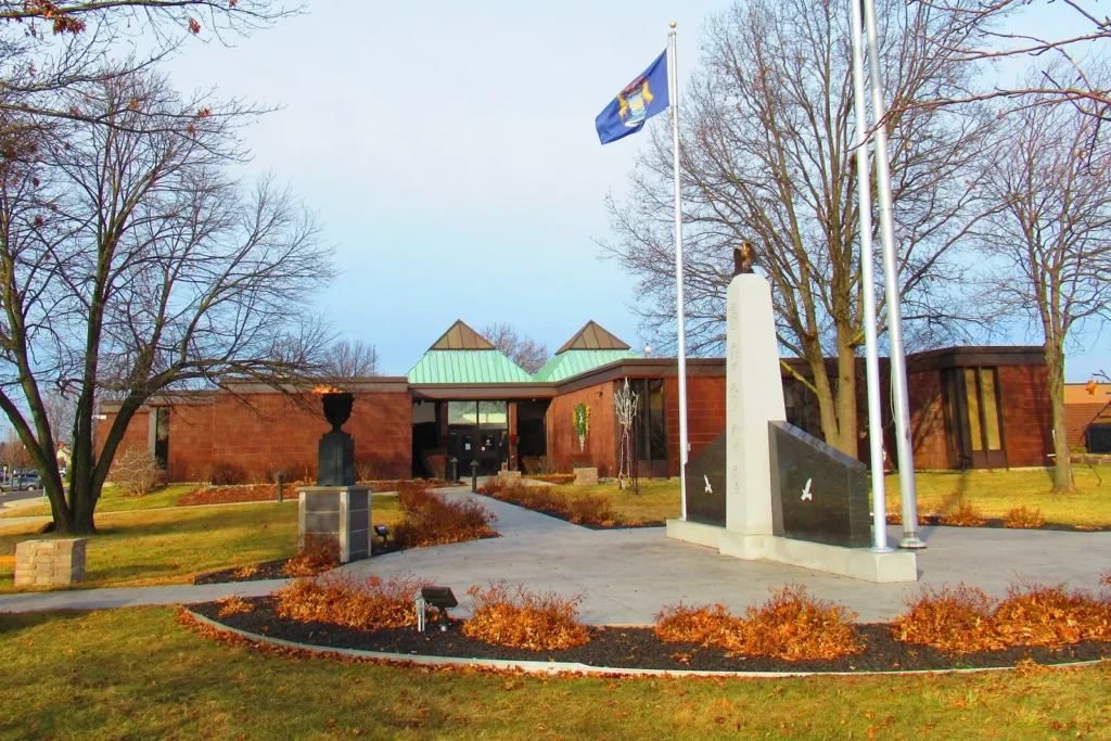 Woodhaven City Hall, a brick building with green roof peaks, behind a memorial plaza and flagpoles in Woodhaven, Michigan